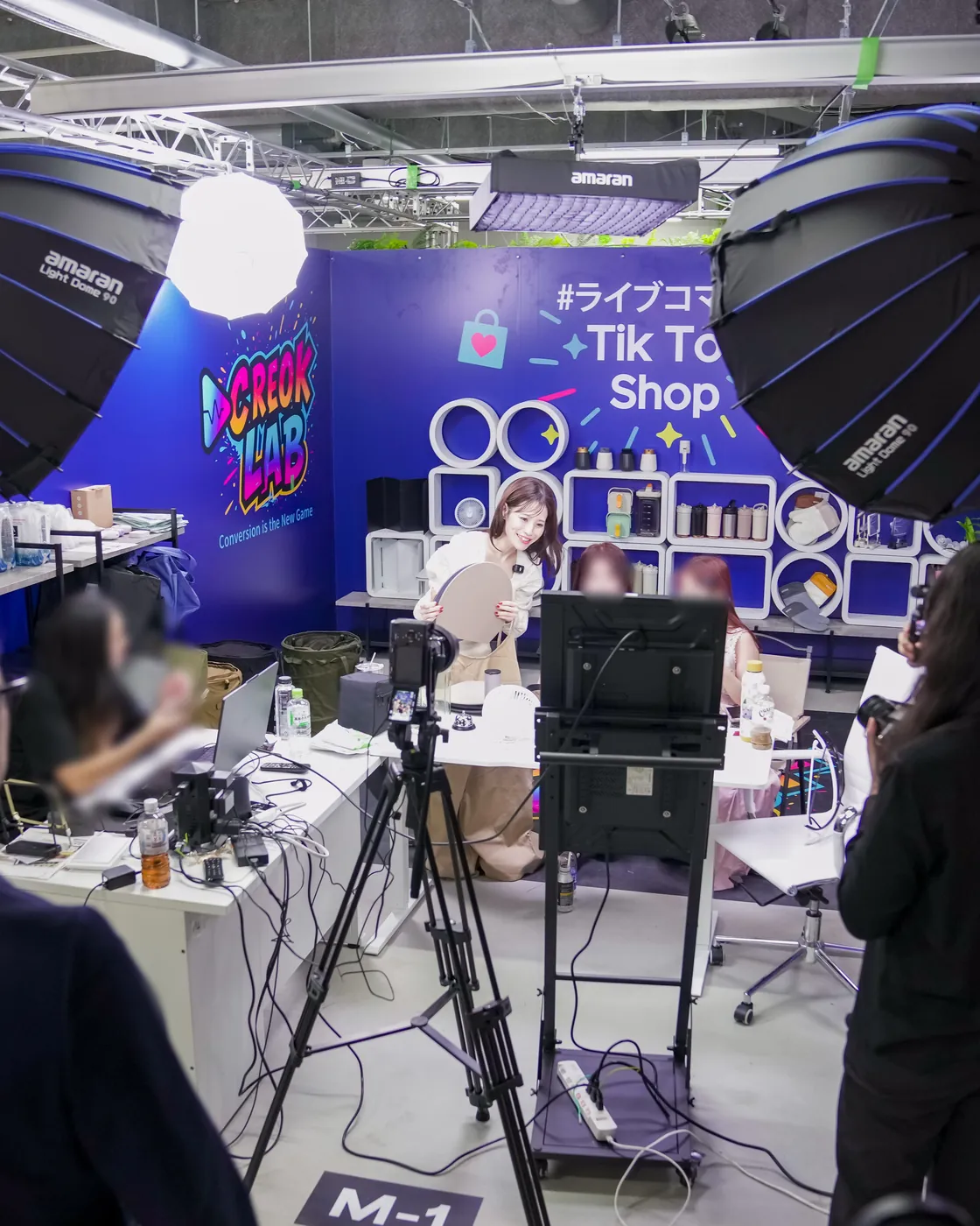 CREOK LAB の個別配信ブース。「CREOK LAB」「#ライブコマース TikTok Shop」の青い壁の前で、照明と三脚のカメラに囲まれてクリエイターが配信している