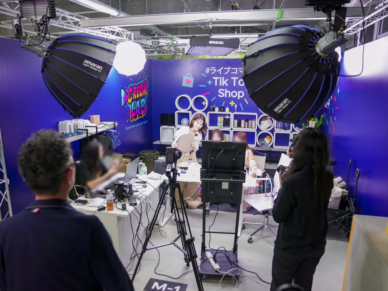 CREOK LAB の個別配信ブース。「CREOK LAB」「#ライブコマース TikTok Shop」の青い壁の前で、照明と三脚のカメラに囲まれてクリエイターが配信している